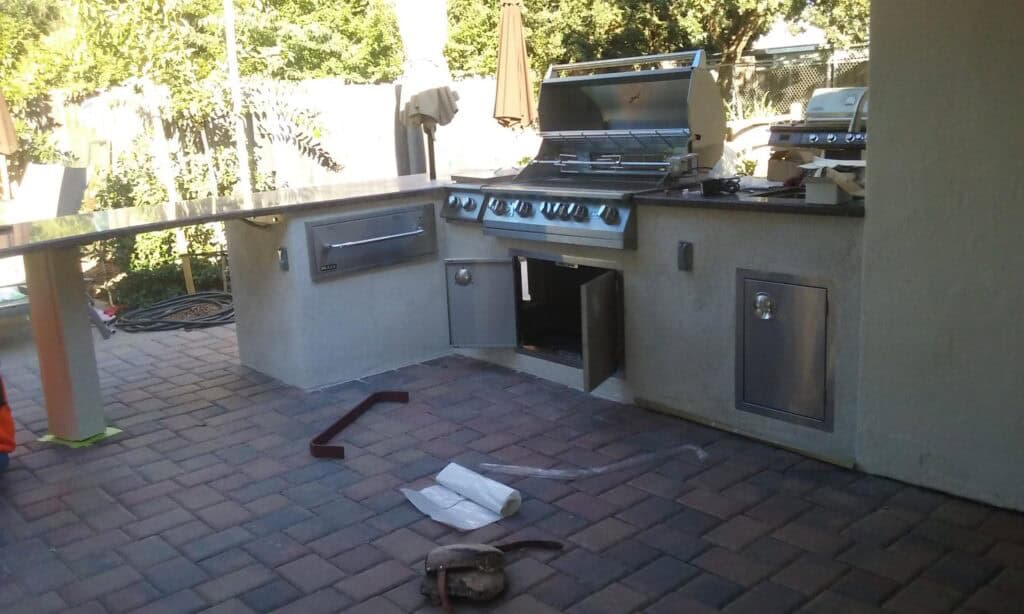 Outdoor kitchen with grill, cabinets, and appliances set on a brick patio in a green backyard.
