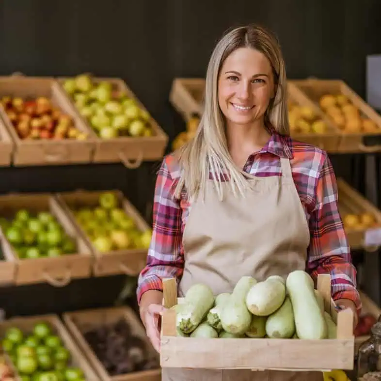 Woman holding a wooden crate of fresh zucchinis at farmers market.