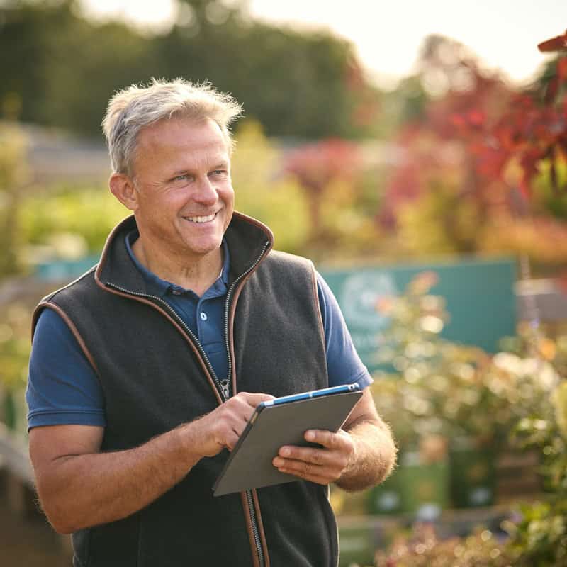 Experienced landscaper smiling outdoors with tablet in hand in a lush garden.