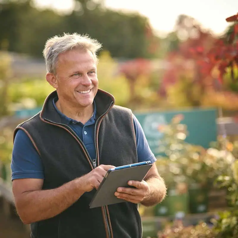 Experienced landscaper smiling outdoors with tablet in hand in a lush garden.